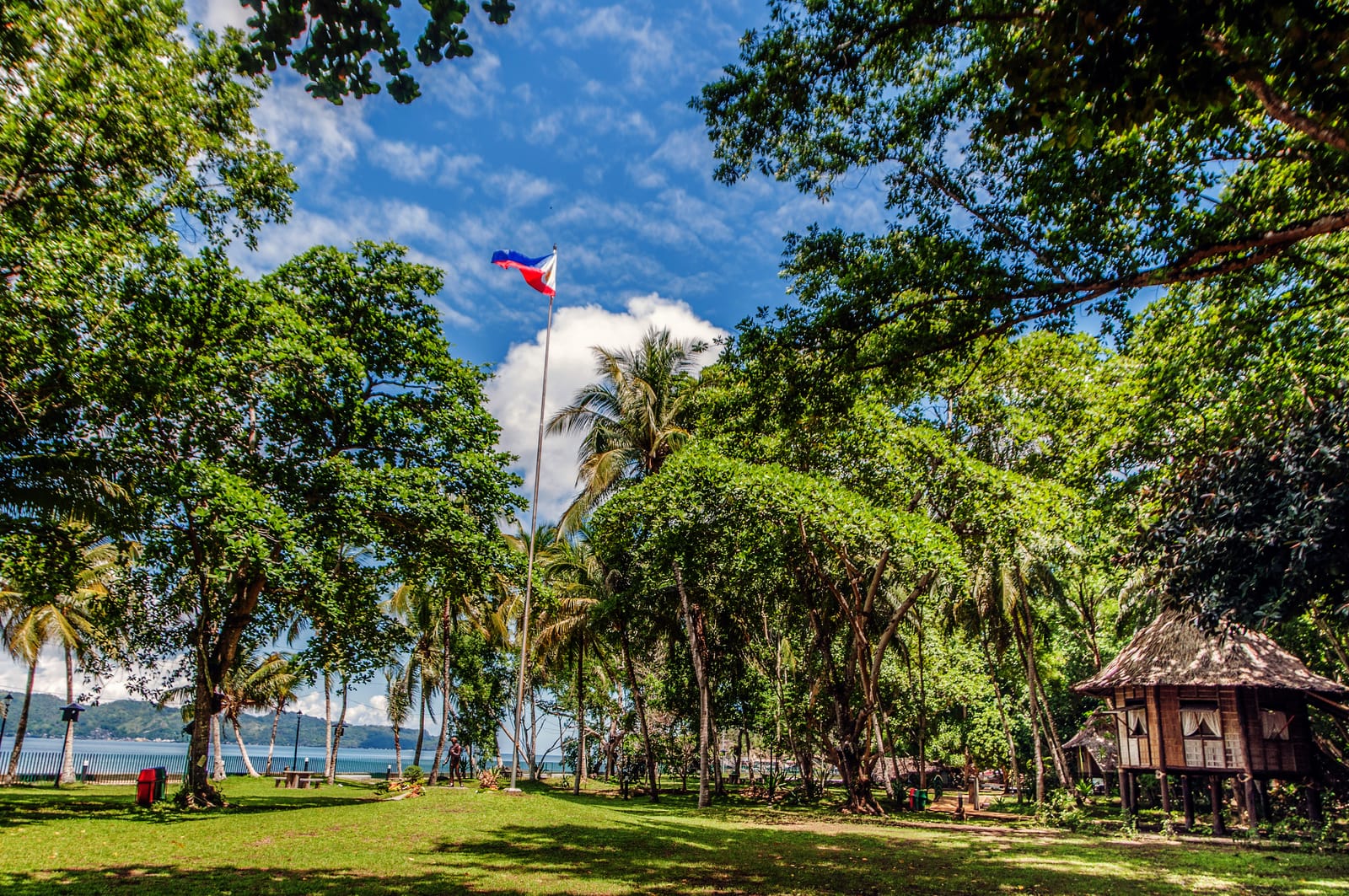 A sunlit lawn under tall shade trees and palms, with the Philippine flag on a high pole, a bamboo-and-thatch house on stilts at the right and the blue bay with green hills beyond.