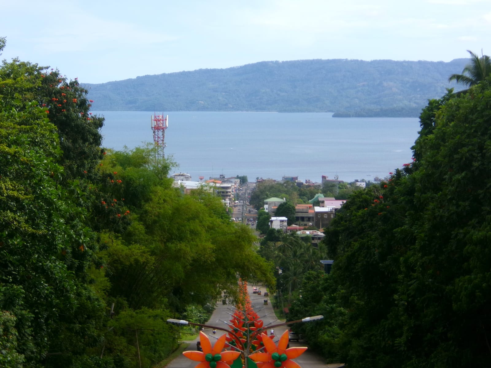 A straight avenue lined with orange flower-shaped lamp decorations running downhill between green trees into a town of rooftops, with a broad grey bay and a long wooded headland beyond.