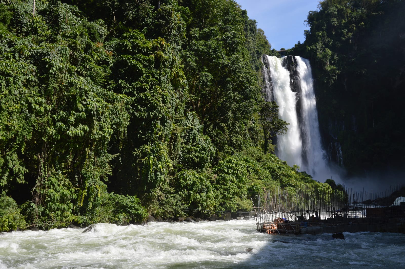 A tall twin waterfall pouring down a dark cliff between forested slopes, above a rushing white river.