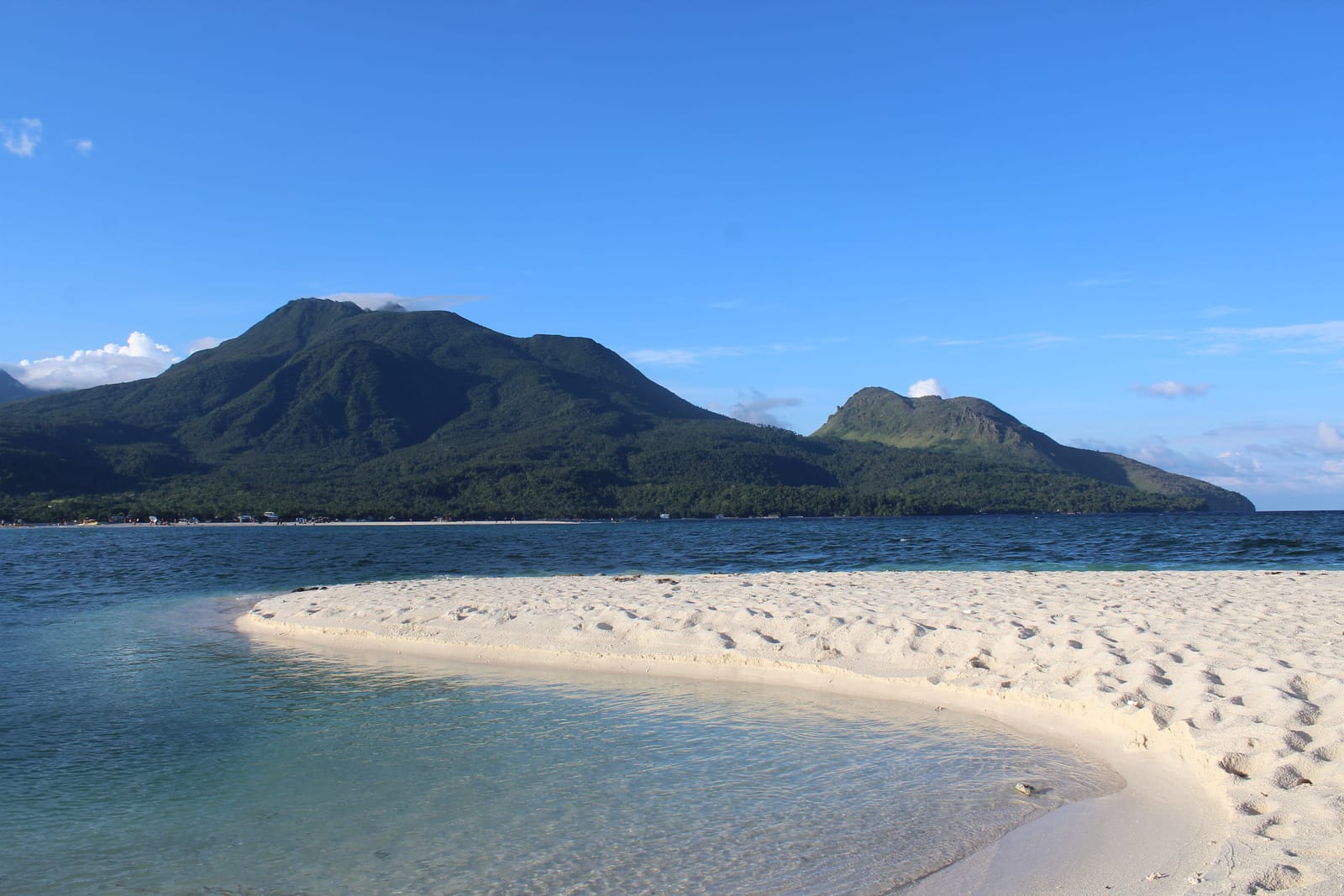 A curving white sandbar in clear shallows, facing a forested volcano with a wisp of cloud at its summit and a smaller rocky peak beside it, under a blue sky.
