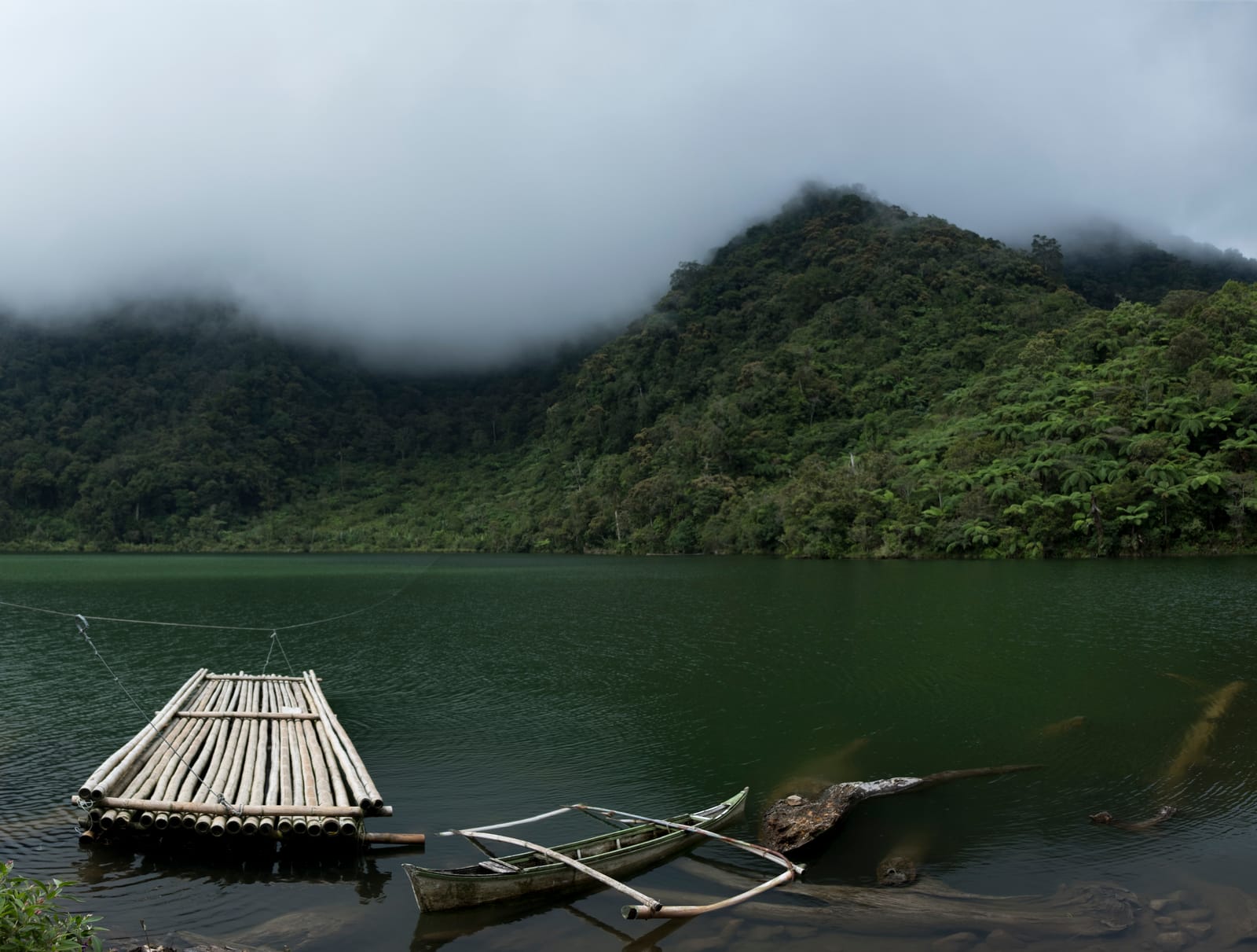 A dark green crater lake ringed by forested slopes under low grey cloud, with a bamboo raft and an outrigger canoe moored at the near shore.