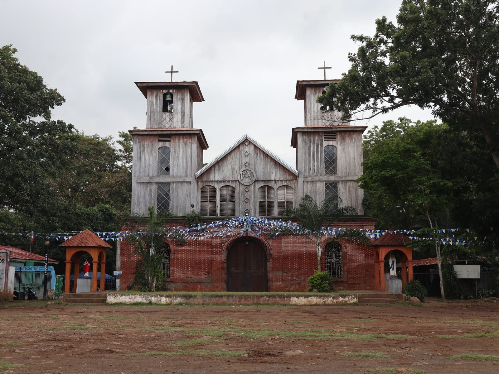 An old church with two weathered wooden bell towers and a wooden gable above a red-brick ground floor with an arched door, hung with blue-and-white bunting, behind a wide dirt plaza under a grey sky.