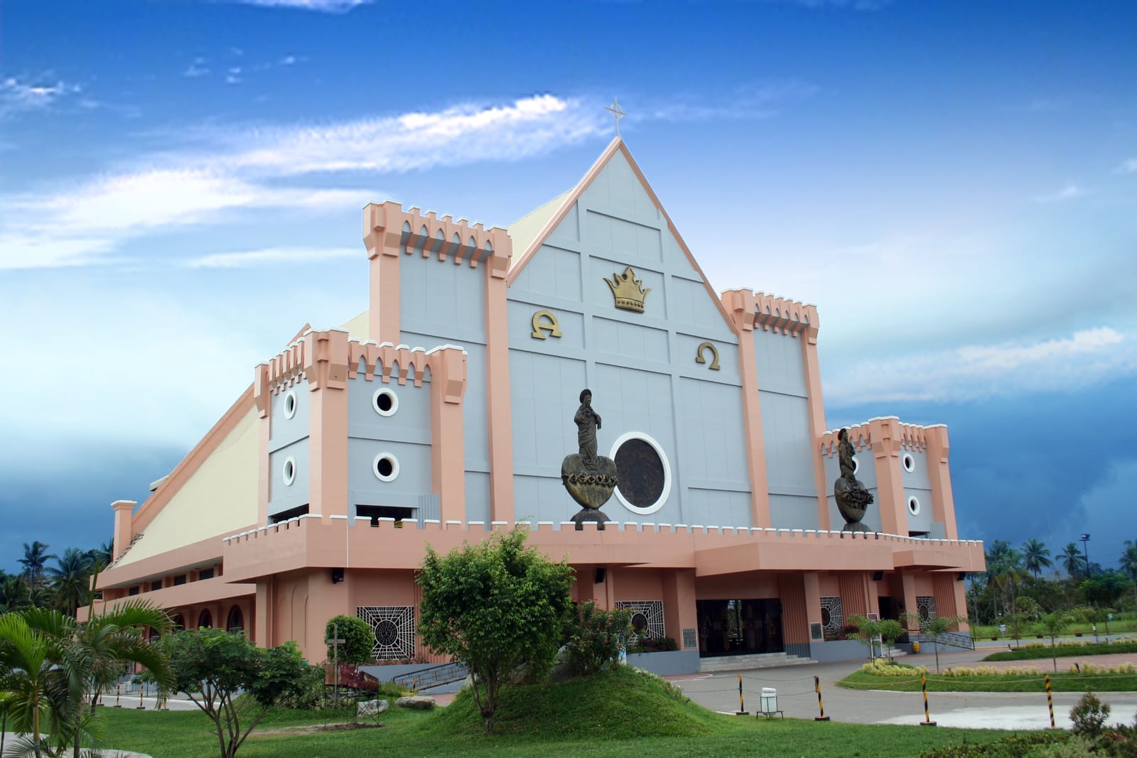A large modern church with a steep grey gable bearing a golden crown and the letters alpha and omega, flanked by pink battlemented towers and bronze statues, behind a grassy mound with shrubs under a blue sky.