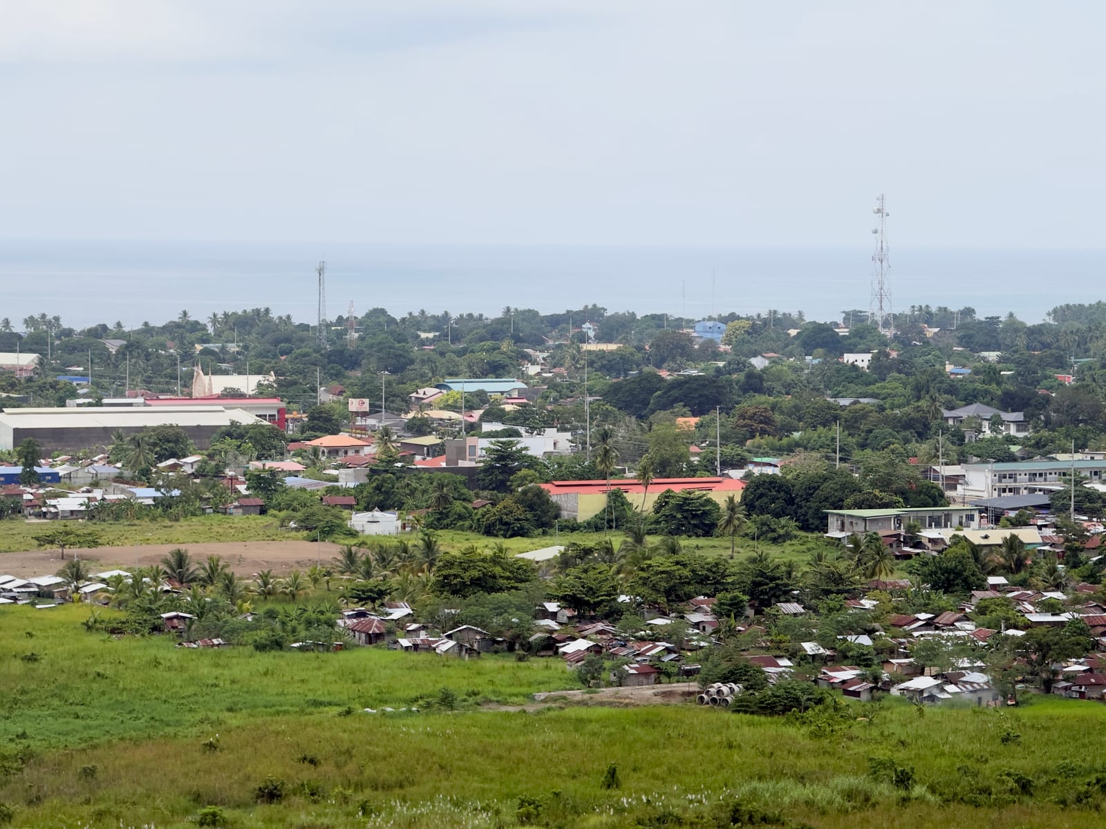 Rooftops of a small coastal town among palms and trees beyond a grassy field, with a long warehouse, a white facade with two pointed spires and two communication towers, and a grey sea under an overcast sky.