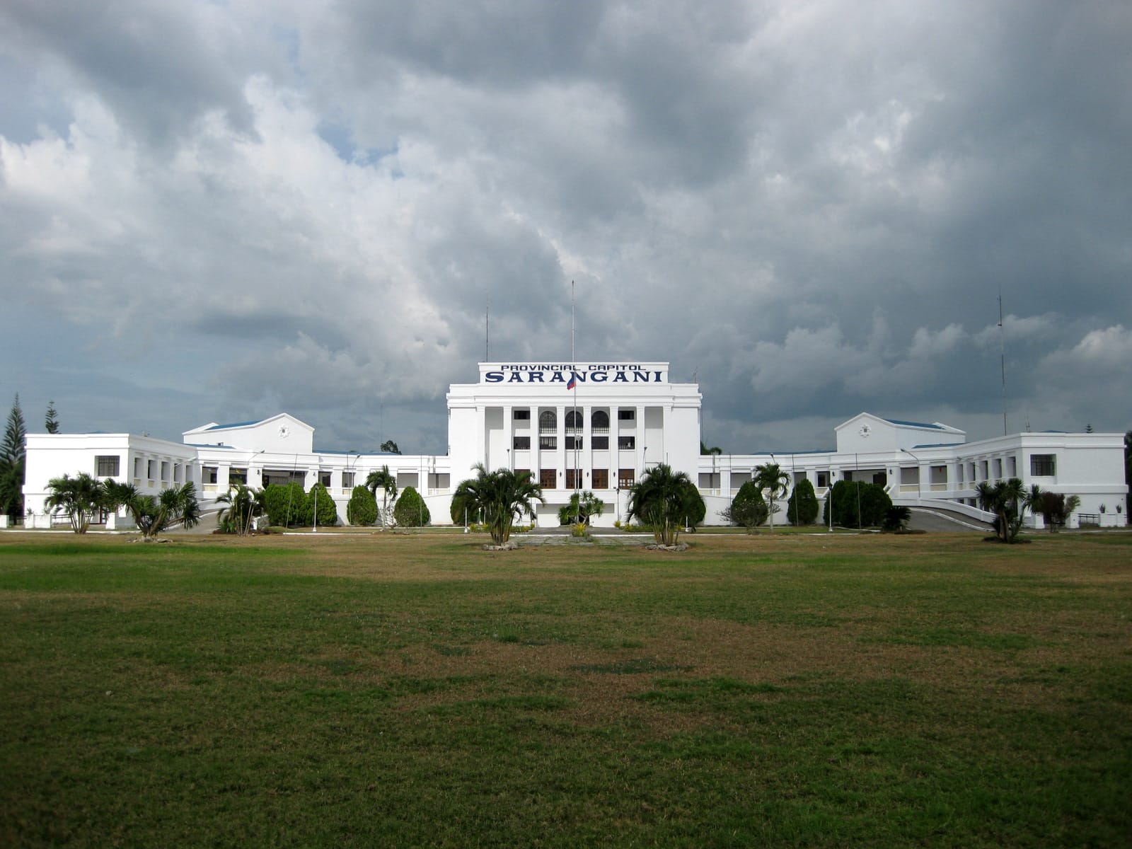 A long white building with a tall central block lettered “Provincial Capitol Sarangani” and low wings to each side, behind palms and a wide lawn, under dark storm clouds.