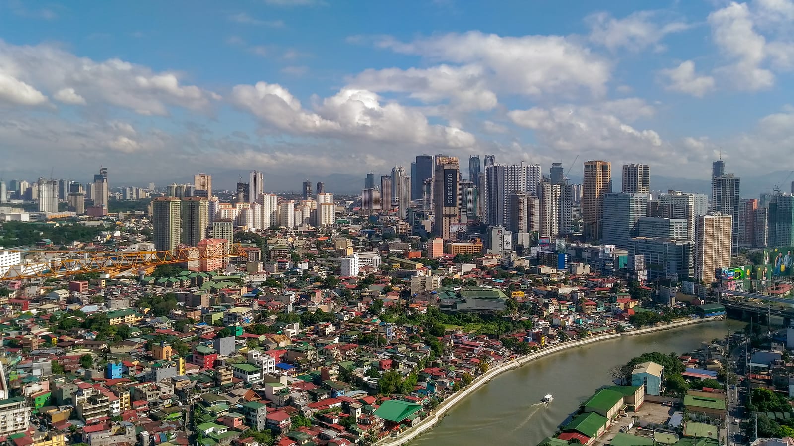 A dense city of low roofs along a curving river, with a cluster of high-rise towers beyond under a blue sky with white clouds.
