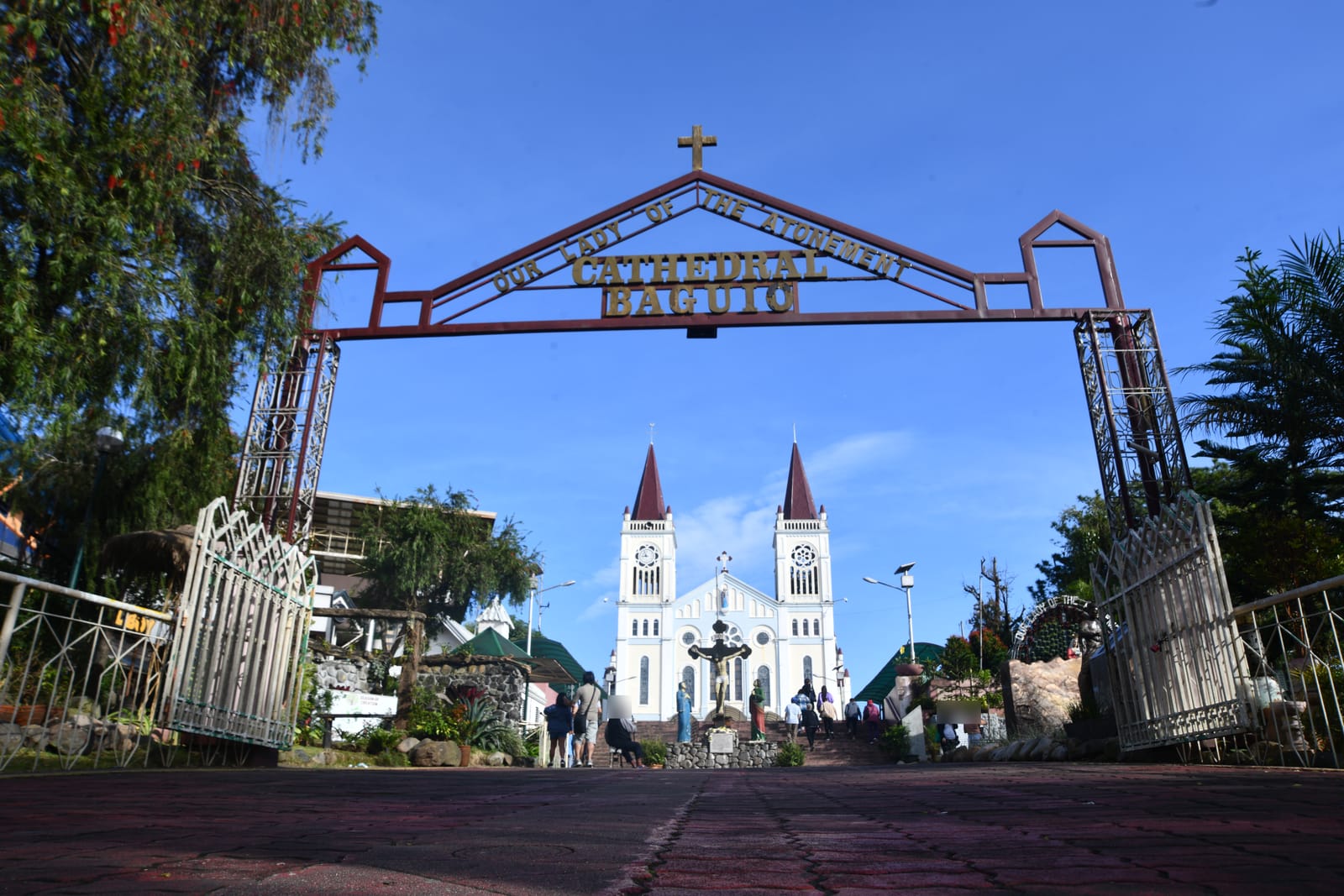 A white church with twin pink spires and rose windows at the top of a stairway, seen through a red iron arch lettered with its name, under a clear blue sky.