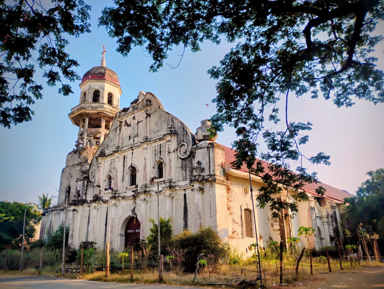 A weathered white baroque church with a scrolled facade, a red roof and a round bell tower capped by a red dome, framed by overhanging branches in early light.