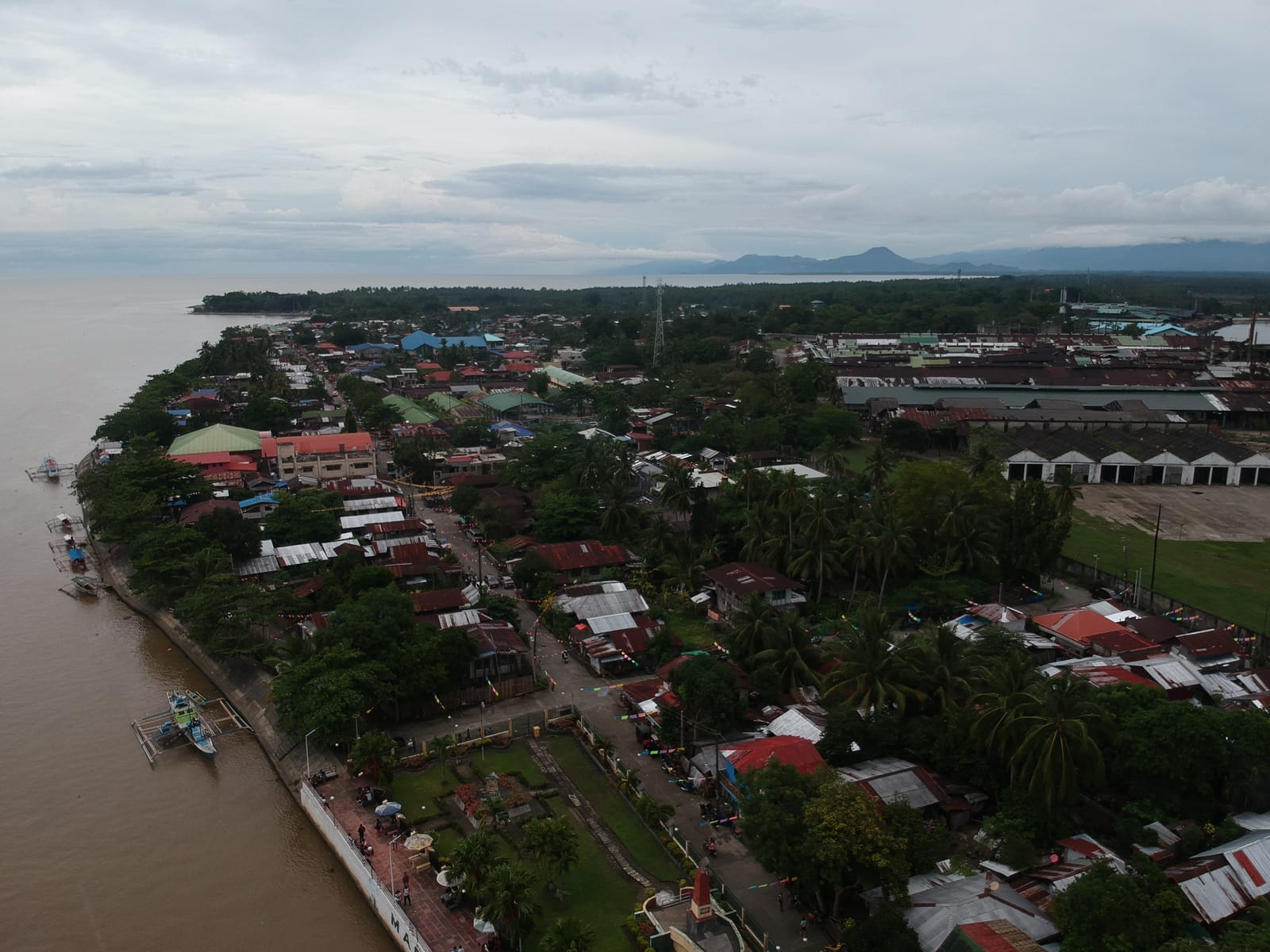 A small town of rusty and red roofs among palms seen from the air beside a wide brown river, with a riverside promenade, a park with a red obelisk, long warehouse roofs and blue mountains on the horizon under a grey sky.
