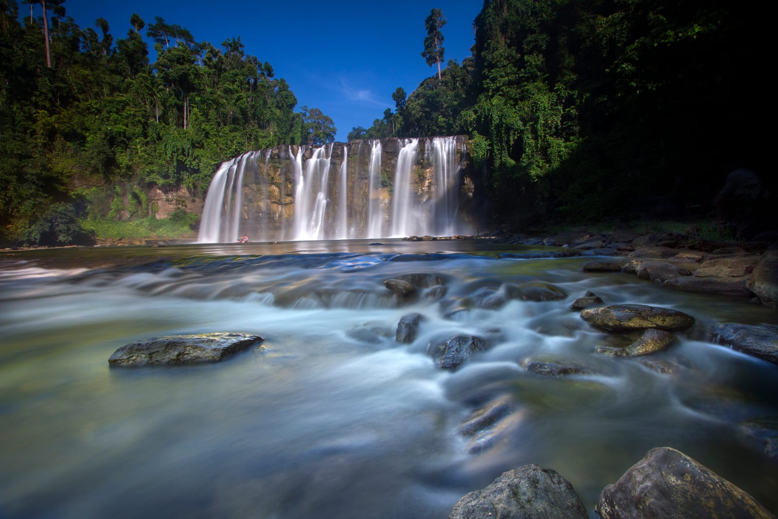 A wide, tiered waterfall in green forest, its water blurred to silk as it runs over rocks in the foreground, under a blue sky.