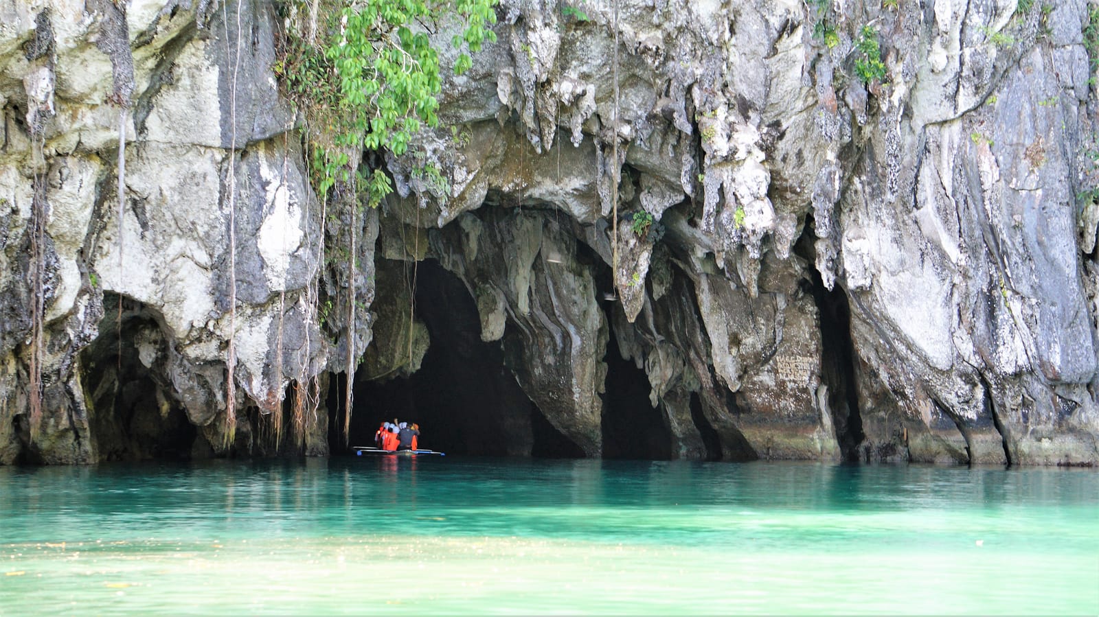 A dark cave mouth in a grey limestone cliff hung with vines, opening onto still turquoise water where a small boat waits.