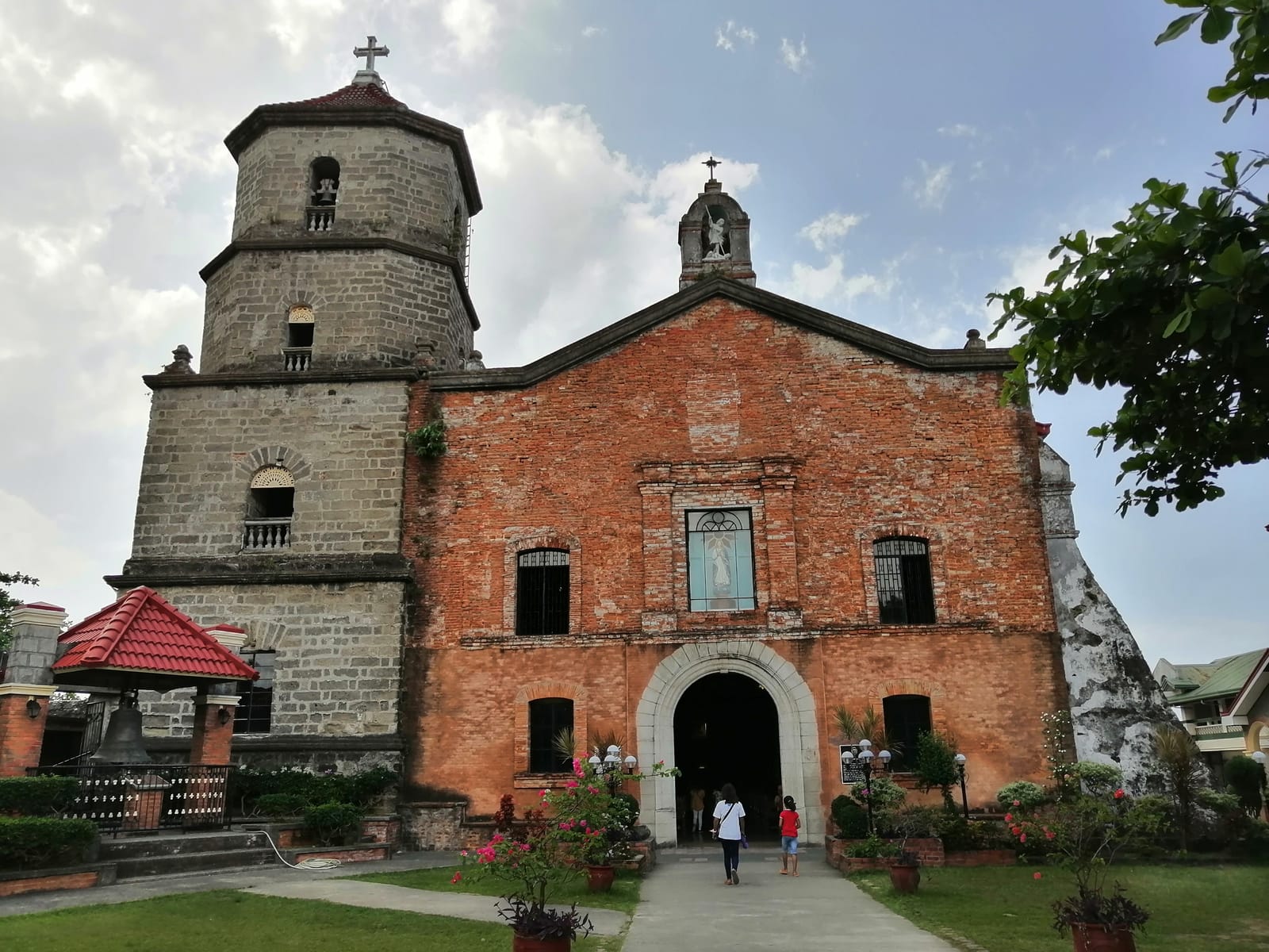A red-brick church facade with a niche above the arched door, beside a squat grey stone bell tower, behind a garden walk under a cloudy sky.