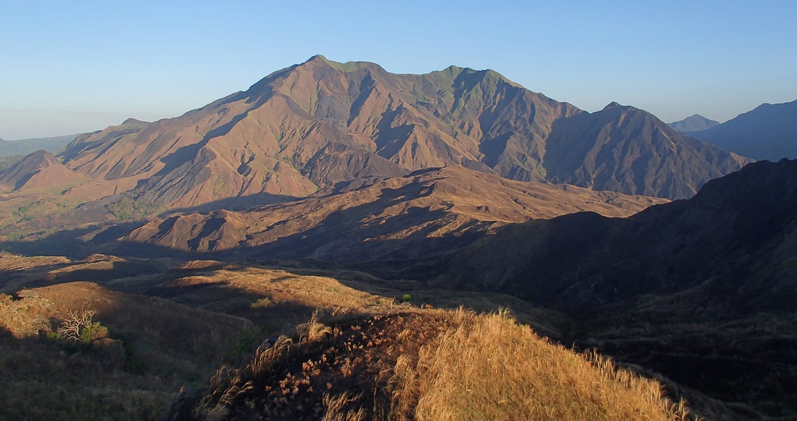 A broad, bare-sloped mountain glowing in low sun above folded brown ridges, with dry golden grass in the foreground under a clear sky.