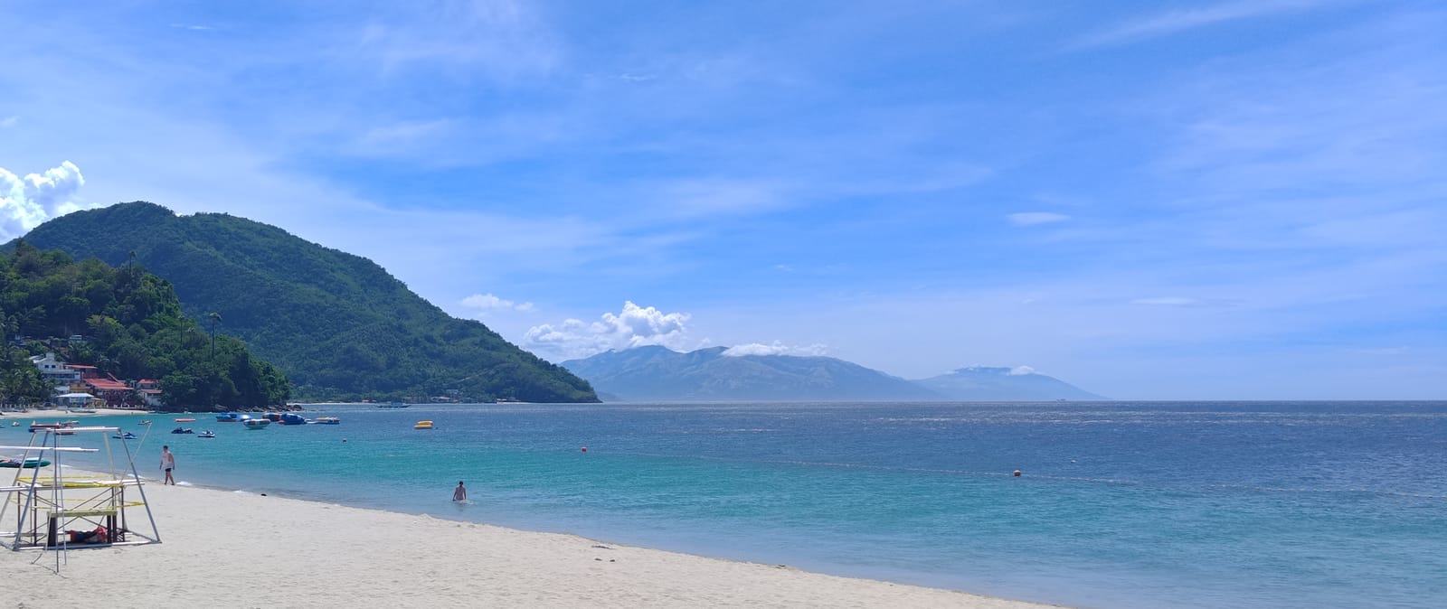 A white-sand beach with a lifeguard stand along clear turquoise water, a green headland at its end and blue mountains across the sea under a bright sky.