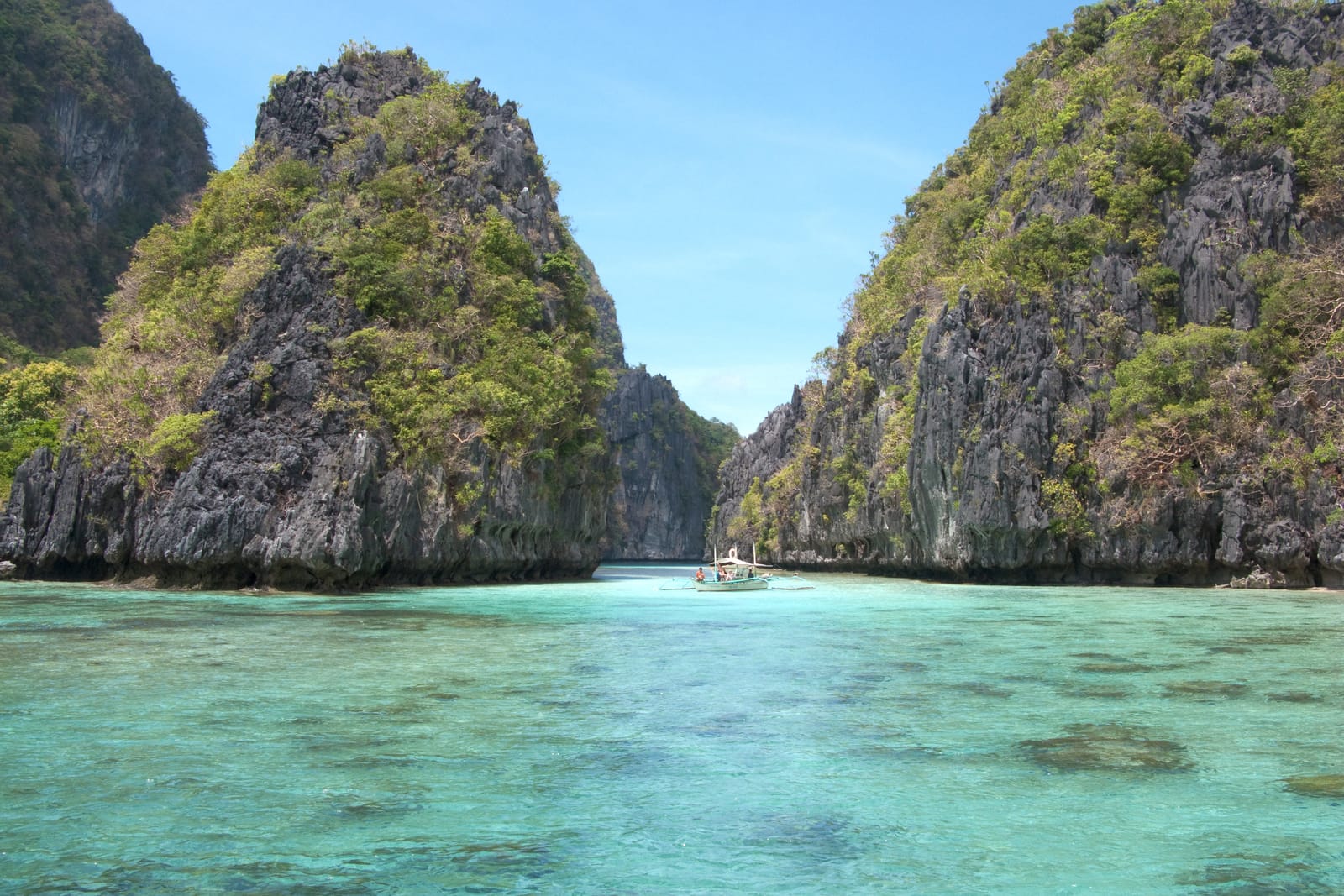 Two steep limestone islands draped in green rising from clear turquoise water, with an outrigger boat in the channel between them.