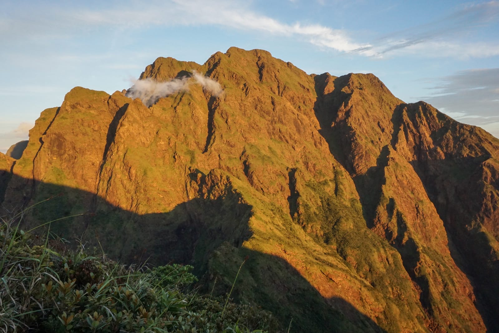 A jagged mountain summit of sheer, grass-streaked ridges lit gold by low sun, with a wisp of cloud on its flank under a pale blue sky.