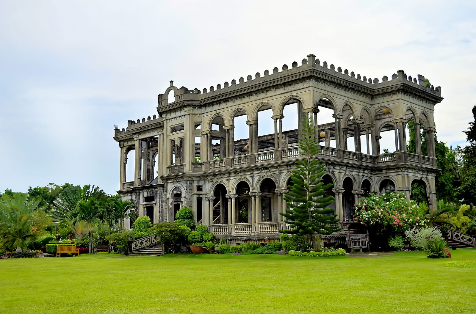 The roofless two-storey shell of a grand mansion with arched windows and balustrades, standing on a wide lawn among palms and flowering shrubs.