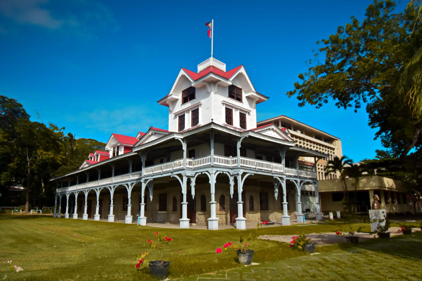 A white wooden hall with red roofs, a tall gabled tower and long verandas on slender white posts, on a lawn under a deep blue sky.