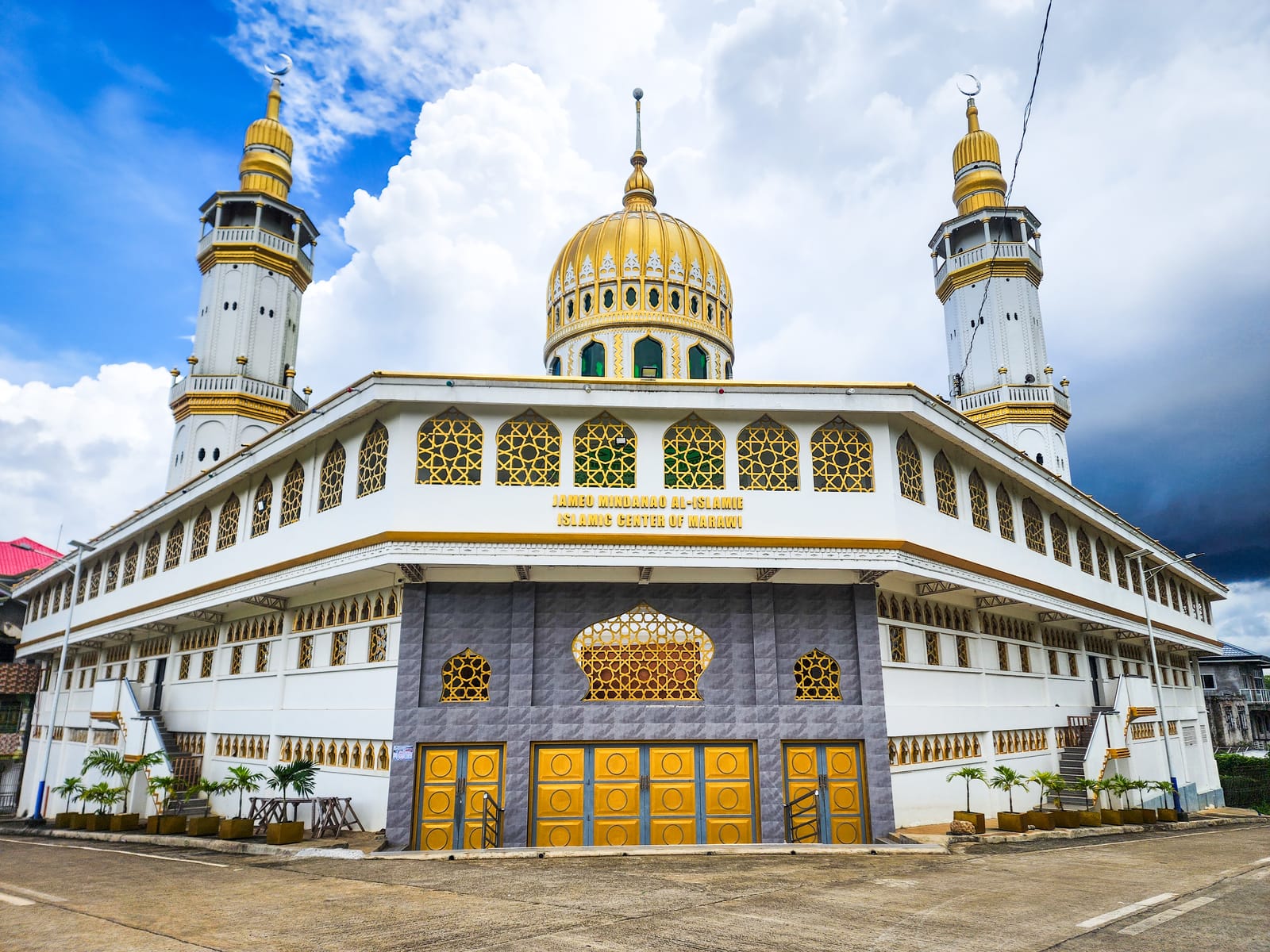 A white mosque with a golden central dome and two tall white-and-gold minarets, its upper walls set with gold lattice windows and lettered with its name, under a blue sky with towering white clouds.