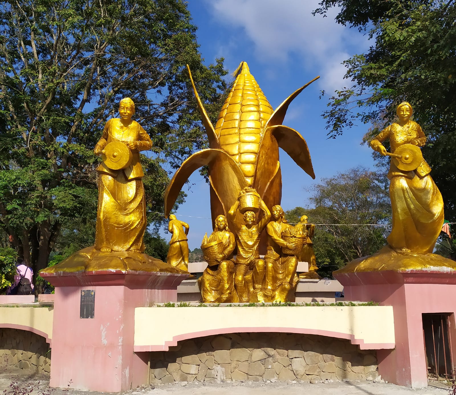 A giant golden ear of corn with gilded farmers carrying baskets at its foot, between two golden statues of women beating gongs on pink pedestals, under trees and a blue sky.